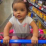 aisle, baby, canned_goods, child, clothing, curious, cute, footwear, grip, grocery_store, indoor, nike_logo, person, pink_bows, retail, shopping, shopping_cart, supermarket, toddler, white_shirt