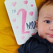 Augustin a rejoint le concours — aidez-le/la à gagner de superbes lots ! baby, infant, face, eyes, skin, clothing, black_shirt, yellow_background, card, text, pink_hearts, milestone, celebration, closeup, portrait, cute, child, young, smile, resting