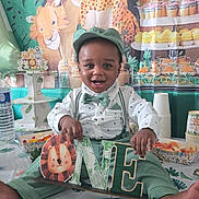 Ibrahim participe au concours pour gagner de l'argent avec cette photo : baby, child, smiling, bow_tie, suspenders, green_outfit, cap, one_sign, birthday_party, jungle_theme, lion_illustration, backdrop, desserts, cupcakes, cups, tablecloth, balloon, decoration_banner, sitting, joyful