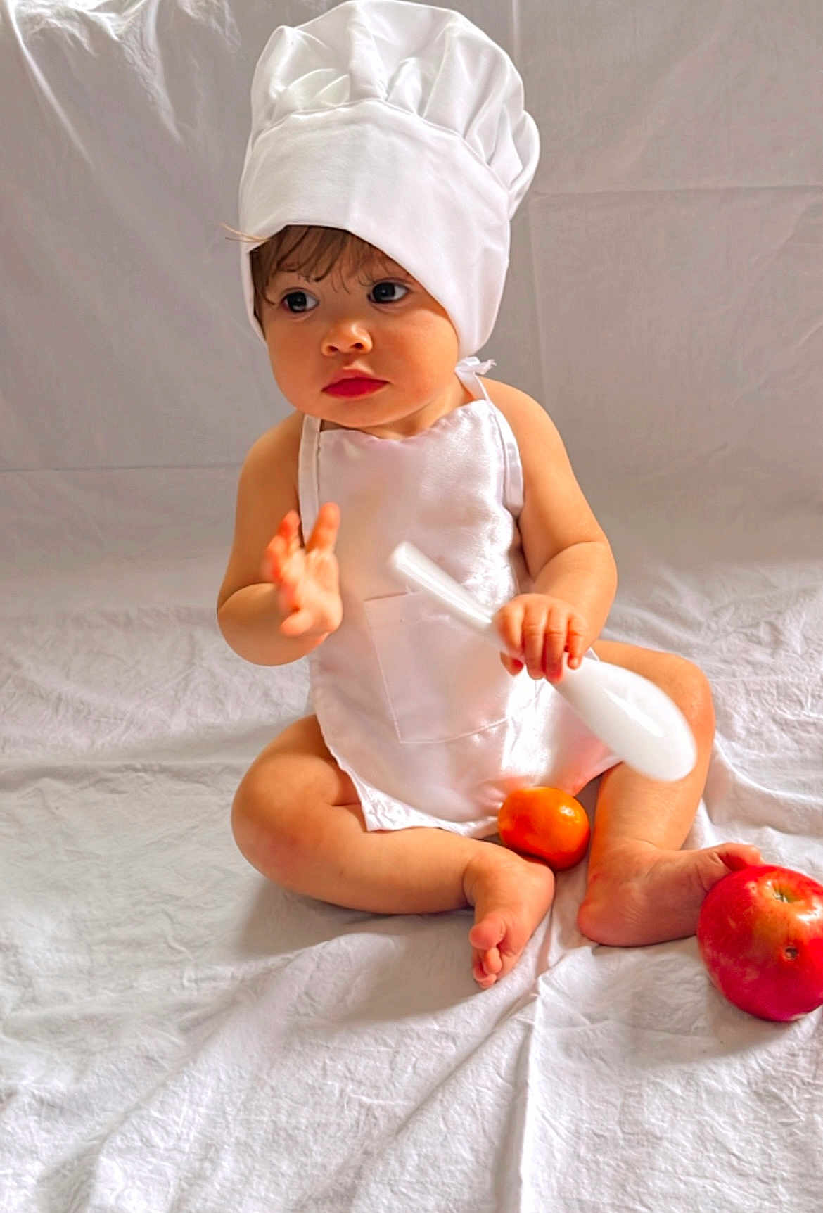 Hellena is registered to the contest to win money with this photo: baby, child, chef_hat, white_apron, sitting, spoon, orange_fruit, apple, barefoot, curious, indoor, white_background, cute, portrait, infant, soft_lighting, food, playful, young_child, closeup