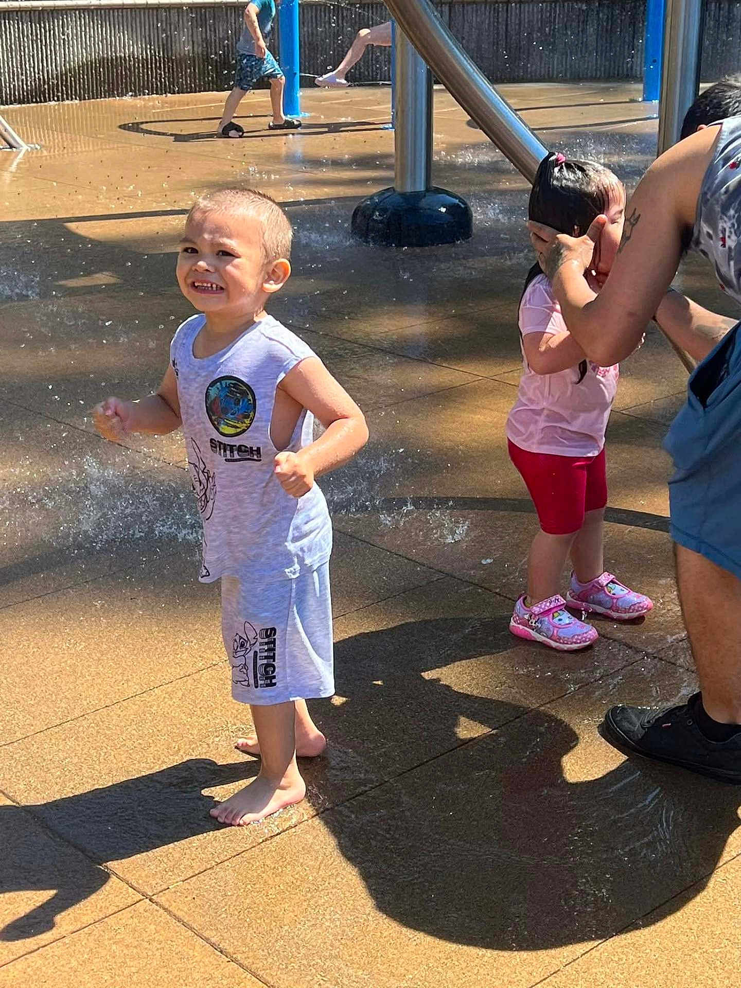 Nathaniel Truncer is registered to the contest to win money with this photo: child, boy, girl, water, splash_pad, playground, outdoor, summer, fun, happy, barefoot, clothing, shoes, water_spray, adult, tattoo, shadow, sunlight, concrete, family