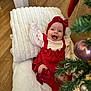 baby, infant, smiling, red_clothing, headband, blanket, christmas_tree, ornaments, holiday, indoor, wood_floor, cushion, happy, cute, festive, decor, person, toddler, celebration, home