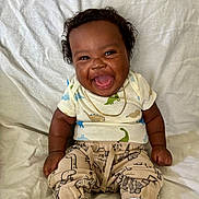 Demari is registered to the contest to win money with this photo: baby, smiling, dinosaur_clothing, white_blanket, sitting, happy, infant, cute, child, portrait, indoors, clothing, socks, gold_necklace, curly_hair, young_child, playful, soft_texture, casual_wear, joy