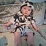 baby, child, infant, couch, cushion, clothing, hat, footwear, sitting, indoor, looking_up, patterned_outfit, plush_furniture, curious, young, person, face, legs, hand, expression