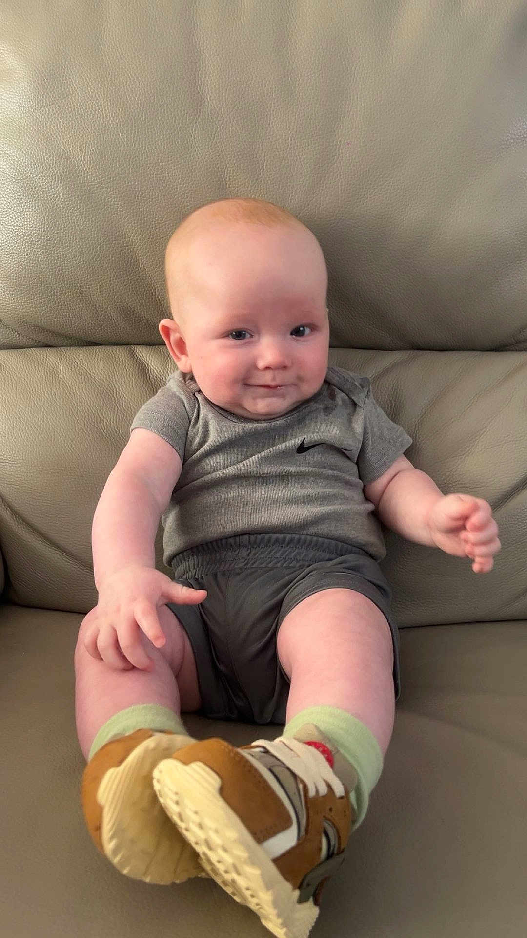 Teddie joined the competition — help win amazing prizes! baby, infant, smiling, sitting, shoe, sneaker, clothing, grey_shirt, shorts, leather_couch, cute, child, person, footwear, happy, indoors, young_child, casual, comfortable, portrait