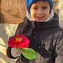 child, smiling, flower, red_flower, blue_hat, winter_coat, gloves, outdoor, sunlight, shadow, wall, leaf, portrait, young_child, happy, nature, seasonal, casual_clothing, person, daylight