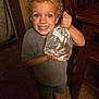 child, boy, curly_hair, smiling, indoor, wooden_floor, plastic_bag, fish, water, holding, casual_clothing, shorts, shoes, happy, portrait, young, person, home, furniture, light