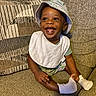 baby, child, smiling, hat, bucket_hat, bib, sock, couch, cushion, indoor, happy, sitting, infant, clothing, footwear, person, portrait, home, cozy, playful
