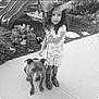 child, dog, outdoor, sidewalk, garden, plant, slide, toy, pinwheel, black_and_white, curly_hair, rain_boots, leash, smile, playful, pet, nature, young_girl, animal, portrait