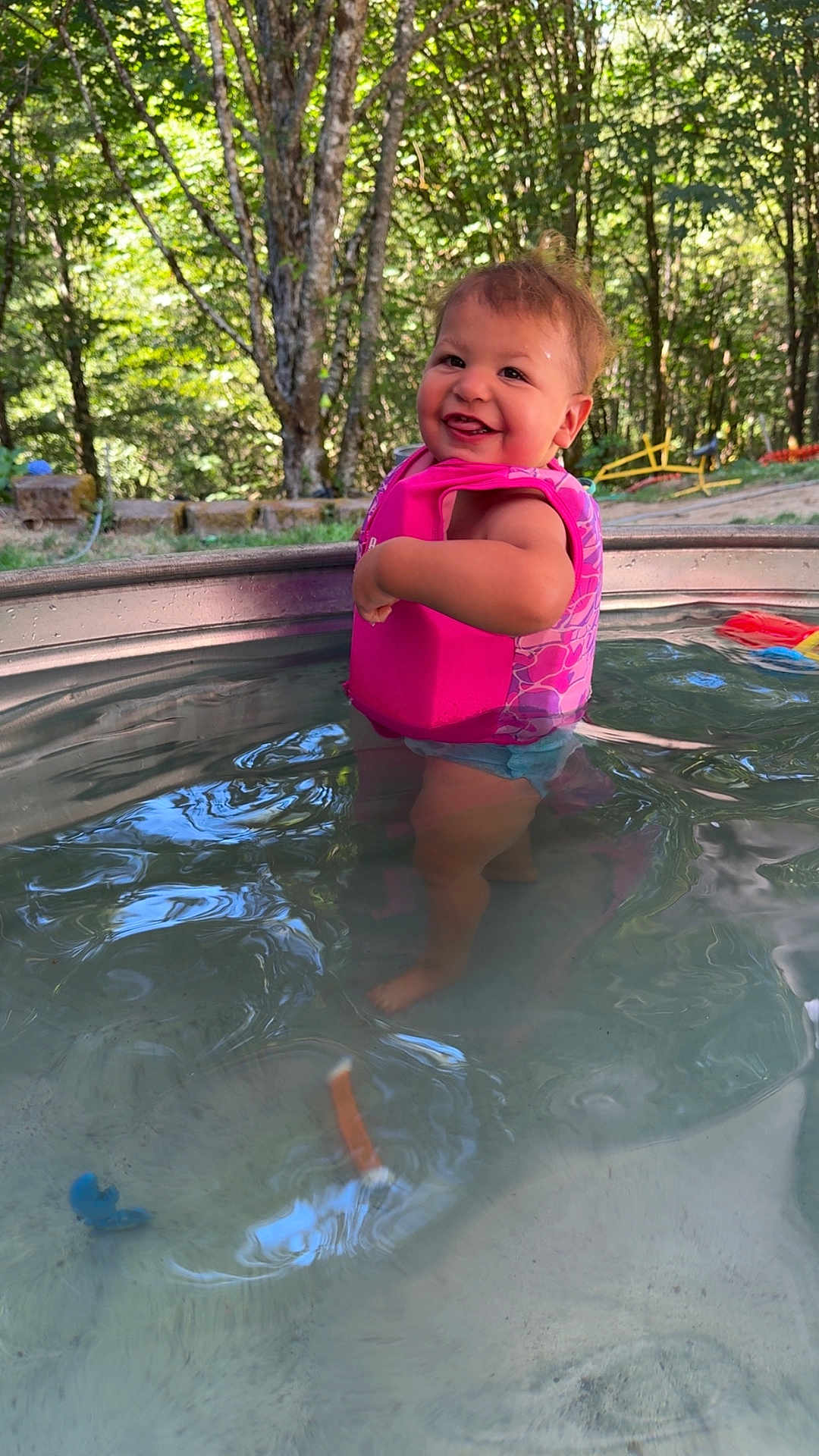 Lillian joined the competition — help win amazing prizes! toddler, child, water, pool, floatation_vest, pink, smiling, outdoor, trees, nature, sunlight, happy, summer, play, barefoot, person, young_child, water_toy, daytime, forest