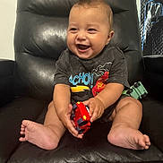 Andres joined the competition — help win amazing prizes! baby, child, smiling, toy_car, black_chair, leather_chair, sitting, happy, indoor, cute, short_hair, barefoot, clothing, playful, infant, person, portrait, furniture, background, mohawk