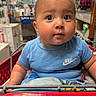Andres is registered to the contest to win money with this photo: baby, child, shopping_cart, blue_shirt, mohawk, face, eyes, store, aisle, shopping, retail, blanket, clothing, person, infant, cute, indoor, head, expression, young