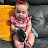 baby, child, red_bow, checkered_dress, couch, orange_pillow, feet, toy, sitting, curious, indoor, person, infant, hair_accessory, plaid, skin, furniture, looking_down, small_hands, expression