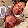 baby, adult_male, father, parent_child, smiling, laughing, cuddle, portrait, close_up, bed, pillow, blanket, white_tshirt, facial_hair, home_interior, wall_decoration, hat, cozy, resting, happy