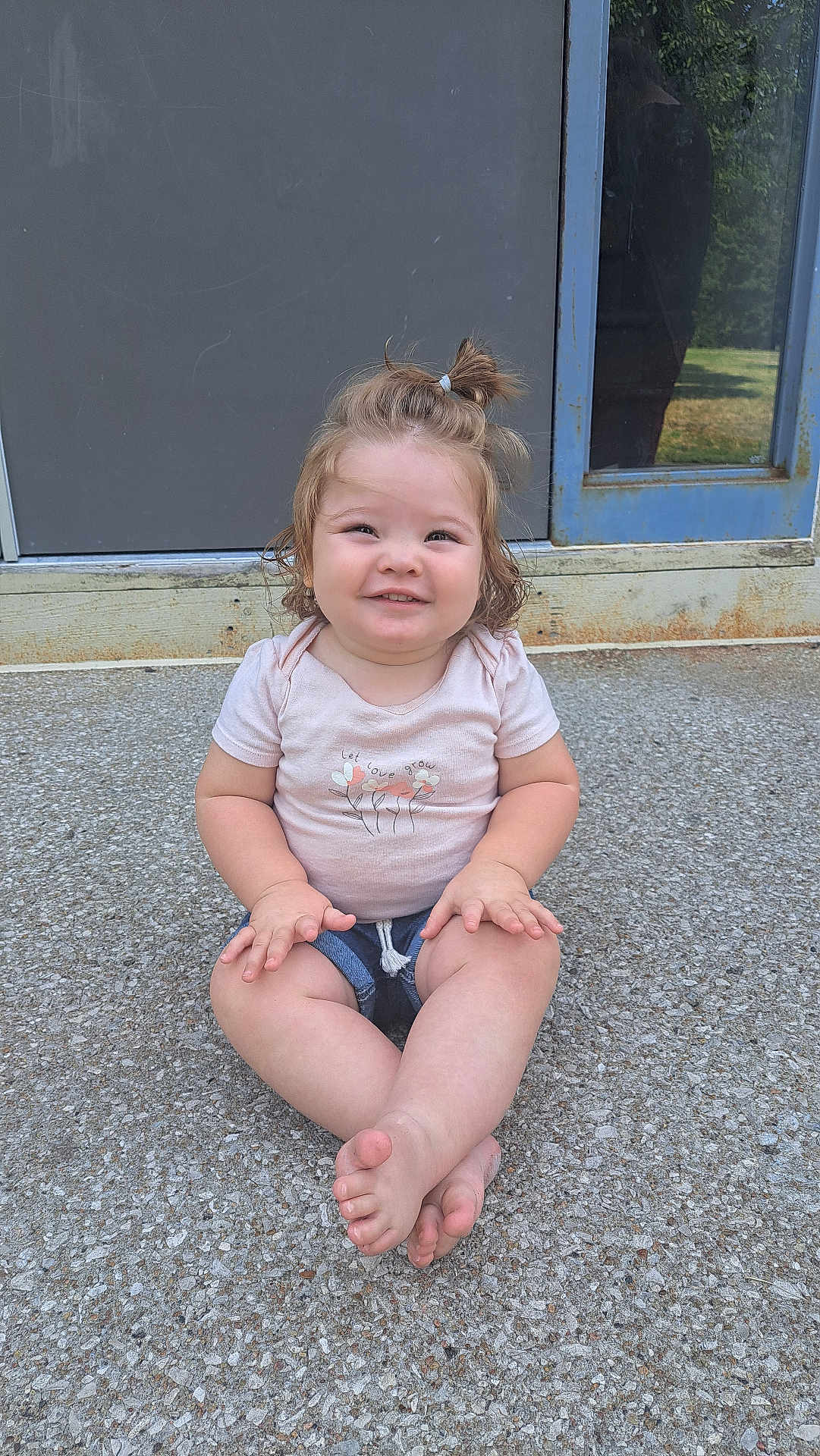 Ember joined the competition — help win amazing prizes! toddler, child, smiling, sitting, cross_legged, ponytail, pink_shirt, denim_shorts, outdoor, door, window, reflection, concrete, gravel, happy, person, casual_clothing, cute, young_child, daylight