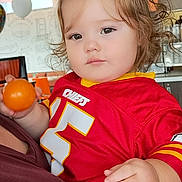 Ember is registered to the contest to win money with this photo: toddler, child, person, indoor, ball, red_jersey, chiefs, curly_hair, holding, casual_clothing, face, expressionless, balloon, background, seated, adult, hand, light, window, portrait