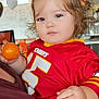 toddler, child, person, indoor, ball, red_jersey, chiefs, curly_hair, holding, casual_clothing, face, expressionless, balloon, background, seated, adult, hand, light, window, portrait