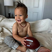 Chance is registered to the contest to win money with this photo: toddler, child, smiling, toy_football, bed, blanket, indoor, happy, bare_chest, necklace, foot, pants, person, cute, hair, playful, home, lamp, furniture, door