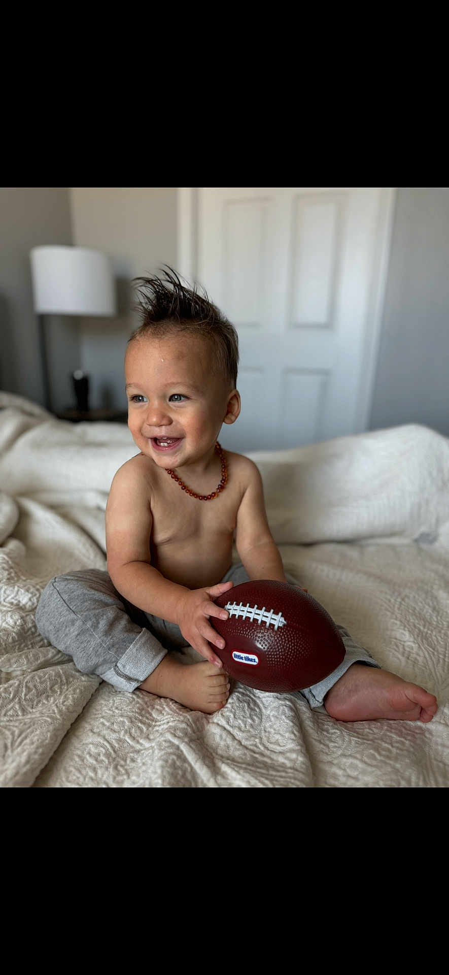 Chance is registered to the contest to win money with this photo: toddler, child, smiling, toy_football, bed, blanket, indoor, happy, bare_chest, necklace, foot, pants, person, cute, hair, playful, home, lamp, furniture, door