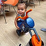 toddler, child, toy, tricycle, indoor, flooring, plastic, orange, blue, chair, bag, play, happy, person, smile, hair, pigtails, furniture, footwear, casual