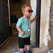 Ysaac participe au concours pour gagner de l'argent avec cette photo : black_shorts, blue_shirt, casual_clothing, child, concrete_floor, doorway, footwear, handrail, indoor, pacifier, pacifier_clip, person, short_hair, sneakers, socks, stairs, standing, sunglasses, toddler, wall