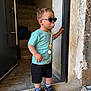 black_shorts, blue_shirt, casual_clothing, child, concrete_floor, doorway, footwear, handrail, indoor, pacifier, pacifier_clip, person, short_hair, sneakers, socks, stairs, standing, sunglasses, toddler, wall
