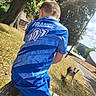 boy, child, dog, football_jersey, france, sportswear, outdoor, park, sunlight, trees, grass, pathway, stone_wall, sitting, back_view, short_hair, blue_clothing, pet, leash, summer