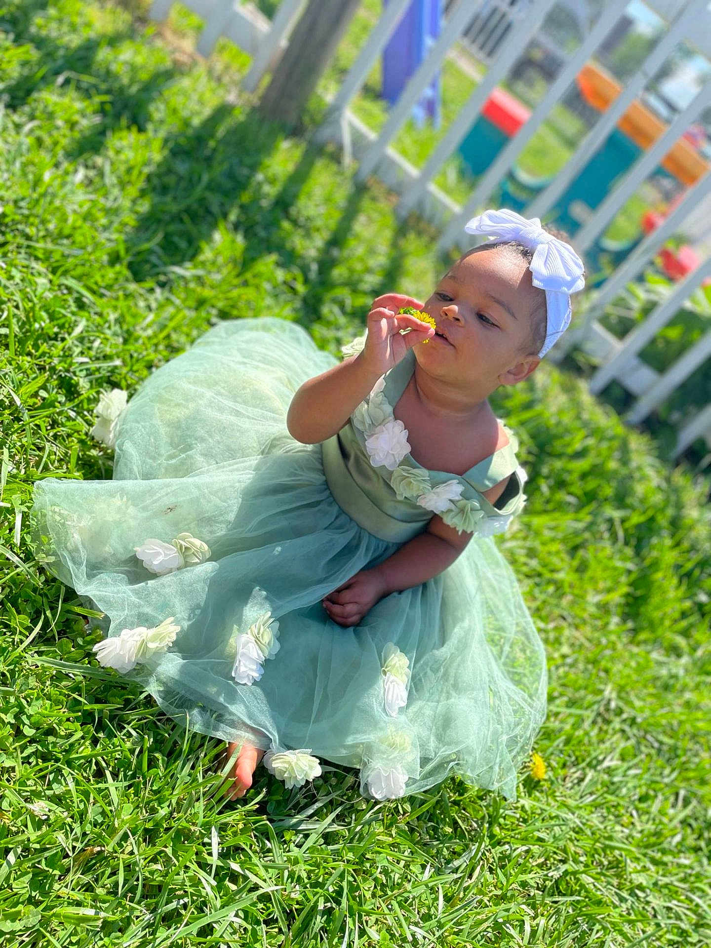 Bessie is registered to the contest to win money with this photo: toddler, child, grass, green_dress, flower, barefoot, outdoor, sunlight, headband, white_flower, picket_fence, playground, curious, sitting, nature, summer, garden, cute, baby, portrait