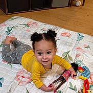 Leighlanni is registered to the contest to win money with this photo: toddler, child, smiling, lying_down, doll, toy, quilt, floral_pattern, yellow_shirt, gray_pants, hair_buns, indoor, wooden_floor, happy, playtime, cute, person, baby, small_toys, floor_mats