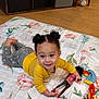 toddler, child, smiling, lying_down, doll, toy, quilt, floral_pattern, yellow_shirt, gray_pants, hair_buns, indoor, wooden_floor, happy, playtime, cute, person, baby, small_toys, floor_mats