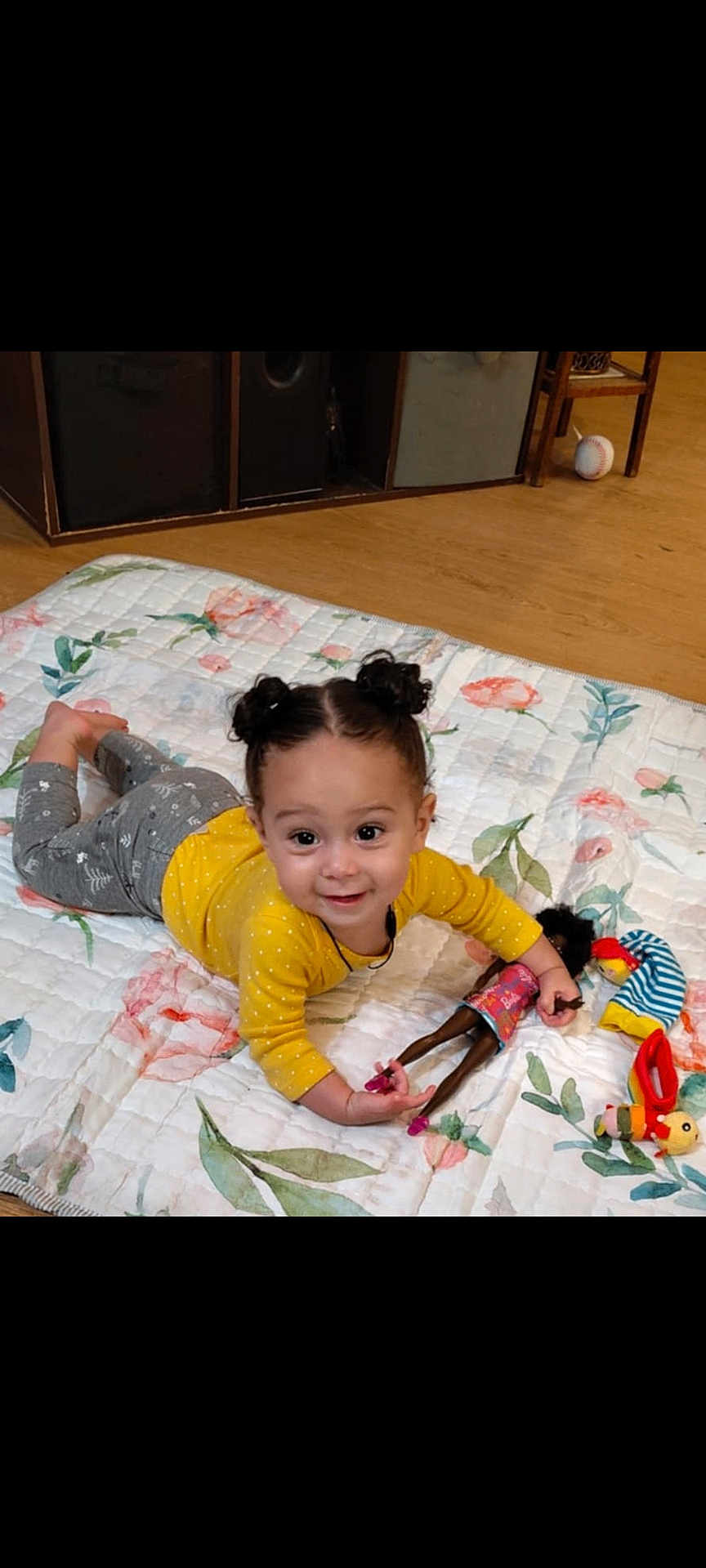 Leighlanni is registered to the contest to win money with this photo: toddler, child, smiling, lying_down, doll, toy, quilt, floral_pattern, yellow_shirt, gray_pants, hair_buns, indoor, wooden_floor, happy, playtime, cute, person, baby, small_toys, floor_mats
