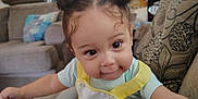 Leighlanni is registered to the contest to win money with this photo: toddler, baby_seat, child, indoors, living_room, curly_hair, double_buns, smiling, reaching, couch, pillows, lamp, window_blinds, casual_clothing, yellow_straps, playful, person, furniture, home_decor, cute