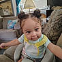 toddler, baby_seat, child, indoors, living_room, curly_hair, double_buns, smiling, reaching, couch, pillows, lamp, window_blinds, casual_clothing, yellow_straps, playful, person, furniture, home_decor, cute