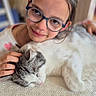 child, glasses, cat, petting, sleeping_cat, tablecloth, indoor, portrait, face, smile, hand, animal, young, person, cozy, closeup, furniture, cute, affection, resting