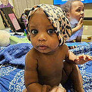 Zaire is registered to the contest to win money with this photo: baby, child, indoor, curious, hand_holding, blanket, patterned, head_wrap, leopard_print, person, young_child, blue, skin, candid, family, bedroom, television, watching, sitting, closeup
