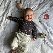 Alba participe au concours pour gagner de l'argent avec cette photo : baby, infant, smiling, lying_down, bed, blanket, toy, bunny_rattle, plush_toy, milestone_card, clothing, white_pants, long_sleeve_shirt, face, hands, feet, indoors, cute, child, happy