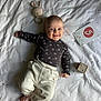 baby, infant, smiling, lying_down, bed, blanket, toy, bunny_rattle, plush_toy, milestone_card, clothing, white_pants, long_sleeve_shirt, face, hands, feet, indoors, cute, child, happy