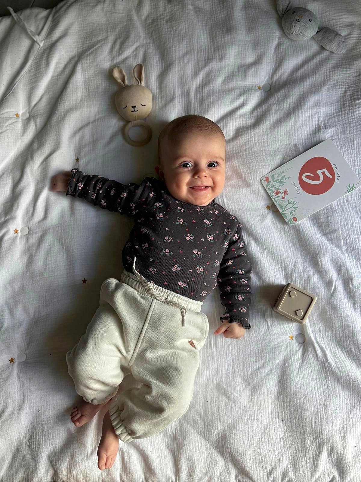 Alba participe au concours pour gagner de l'argent avec cette photo : baby, infant, smiling, lying_down, bed, blanket, toy, bunny_rattle, plush_toy, milestone_card, clothing, white_pants, long_sleeve_shirt, face, hands, feet, indoors, cute, child, happy