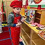 Killian is registered to the contest to win money with this photo: child, chef_hat, red_apron, toy_kitchen, play_area, wooden_shelves, toy_food, colorful_rug, indoor, pretend_play, young_boy, casual_clothing, playroom, sneakers, imaginative_play, holding_object, decor, pizza_mural, focused, educational_toys
