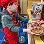 child, toy, pizza, red_hat, red_apron, play_kitchen, carpet, colorful, indoor, pretend_play, hoodie, sneakers, wooden_furniture, cup, oven_mitt, learning_environment, preschool, playroom, young_child, imaginative_play