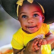 Imani is registered to the contest to win money with this photo: adult_hand, baby, child, close_up, colorful, eyes, face, family, hands, happy, indoor, large_hat, looking_at_camera, photography, portrait, smiling, soft_focus, sun_hat, toddler, yellow_outfit