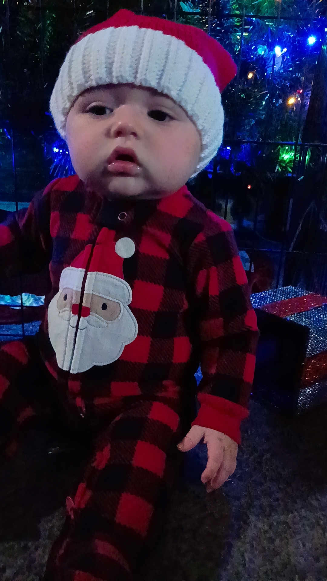 Ezra is registered to the contest to win money with this photo: baby, child, holiday, christmas, santa_hat, knit_hat, red_clothing, plaid, onesie, festive, lights, decorations, indoors, cute, chubby_cheeks, face, hand, sitting, winter, seasonal