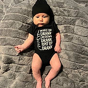 Kane is registered to the contest to win money with this photo: baby, infant, black_onesie, black_hat, blanket, gray_blanket, lying_down, baby_clothing, text_on_clothing, humor, cute, indoors, child, newborn, soft_texture, skin, feet, arms, face, expression