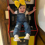 Sofía Zoé joined the competition — help win amazing prizes! baby, child, toddler, smiling, face, portrait, yellow_bow, denim_overalls, floral_shirt, sneakers, sitting, arcade, arcade_machine, pac_man, video_game, indoor, playroom, colorful, happy, gaming