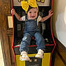 baby, child, toddler, smiling, face, portrait, yellow_bow, denim_overalls, floral_shirt, sneakers, sitting, arcade, arcade_machine, pac_man, video_game, indoor, playroom, colorful, happy, gaming