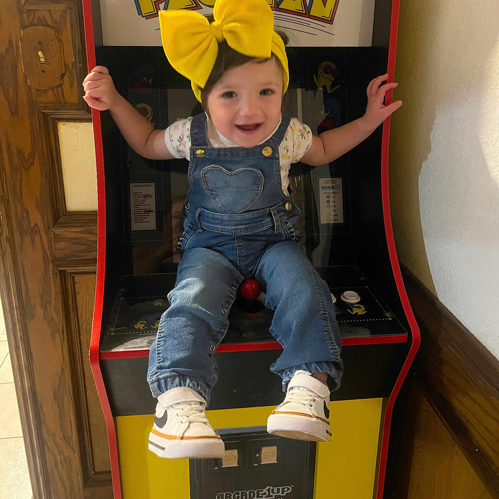 Sofía Zoé joined the competition — help win amazing prizes! baby, child, toddler, smiling, face, portrait, yellow_bow, denim_overalls, floral_shirt, sneakers, sitting, arcade, arcade_machine, pac_man, video_game, indoor, playroom, colorful, happy, gaming
