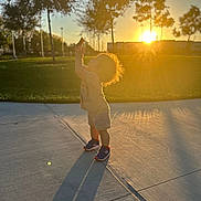 James is registered to the contest to win money with this photo: casual_clothing, child, evening, grass, happy, holding_food, nature, outdoor, park, playful, portrait, shadow, sidewalk, silhouette, sneakers, sunlight, sunset, toddler, trees, young_child