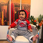Kael a rejoint le concours — aidez-le/la à gagner de superbes lots ! toddler, child, smiling, hoodie, red_clothing, grey_pants, barefoot, chair, interior, wooden_floor, artwork, cityscape, flower_arrangement, plant, happy, indoor, person, furniture, portrait, casual_clothing
