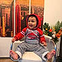 toddler, child, smiling, hoodie, red_clothing, grey_pants, barefoot, chair, interior, wooden_floor, artwork, cityscape, flower_arrangement, plant, happy, indoor, person, furniture, portrait, casual_clothing