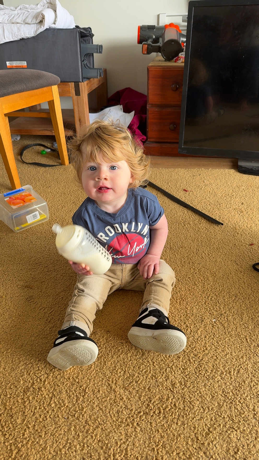Lane joined the competition — help win amazing prizes! toddler, child, milk_bottle, carpet, indoor, sitting, blue_tshirt, pants, shoes, blonde_hair, furniture, drawer, television, chair, toy_box, face, person, home, floor, casual